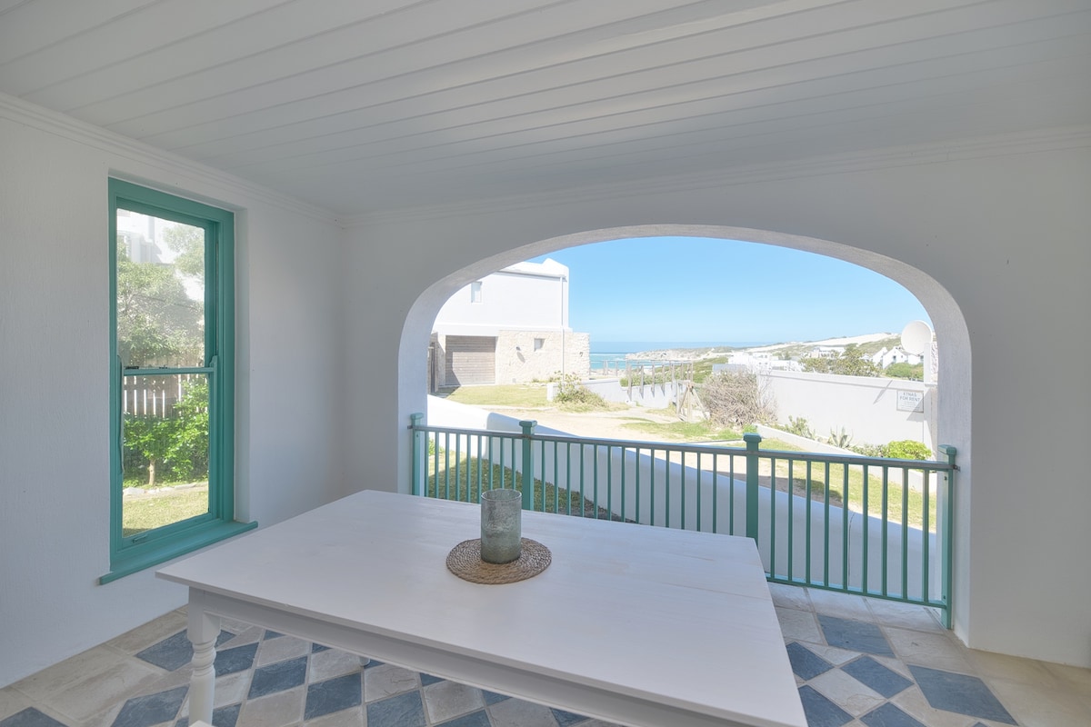 An outdoor dining area is featured under a covered patio, showcasing a white table with a decorative centerpiece. The space is framed by a coastal view visible through an archway, with grass and greenery beyond, complementing the bright blue sky.