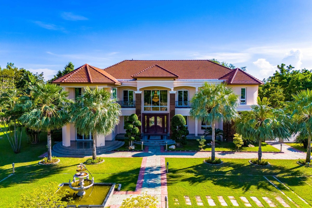 The grand mansion is framed by lush greenery and features a striking entrance. Palm trees and well-manicured shrubs line the walkway, leading to the elegant facade with large windows and a decorative entryway. A serene fountain adds to the tranquil atmosphere of the expansive garden.