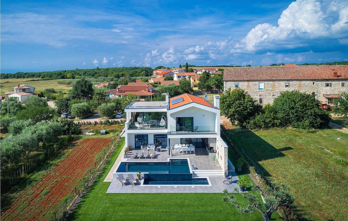 An aerial view of Villa Terani highlights its modern architecture and spacious layout. The inviting infinity pool is set against a backdrop of lush green areas, while the upper terrace features comfortable seating. Nearby, the vibrant landscape is framed by charming rural structures.