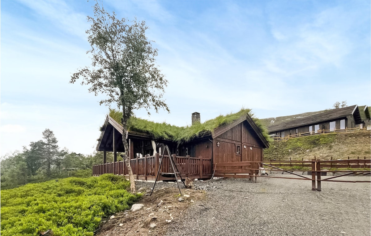 A wooden vacation home with a grass-covered roof is set against a backdrop of natural greenery. The structure features a spacious deck and is surrounded by trees and shrubs, providing a rustic charm in a tranquil outdoor setting.