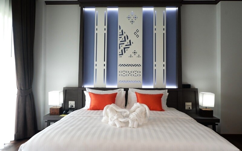 A spacious bed is centered in the room, framed by decorative wall panels featuring intricate patterns. Plush white linens are complemented by vibrant orange pillows. Soft lighting emanates from bedside lamps, creating a calming ambiance. A neatly arranged set of towels adds a touch of elegance.