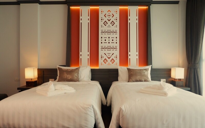 A well-appointed guest room features two spacious beds adorned with white linens. Soft lighting enhances the intricate wall art and the soothing color palette. Bedside lamps provide additional illumination, while curtains frame the windows, allowing natural light to fill the space.