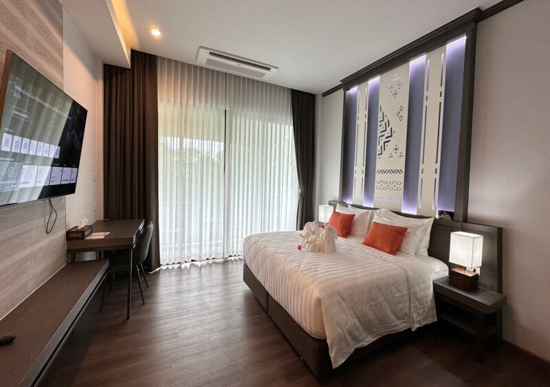 A modern guest room features a spacious bed adorned with striped pillows and a decorative towel arrangement. Natural light floods in through large curtains, complemented by subtle wall artwork and a sleek desk. A flat-screen TV is mounted on the wall, enhancing the contemporary decor.