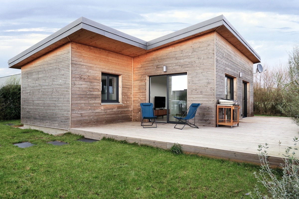A modern house features a wooden exterior and a spacious terrace. Two blue chairs are positioned on the terrace, overlooking a grassy area. Large glass doors allow for natural light, and a dining table is visible on the terrace, providing an inviting outdoor space.