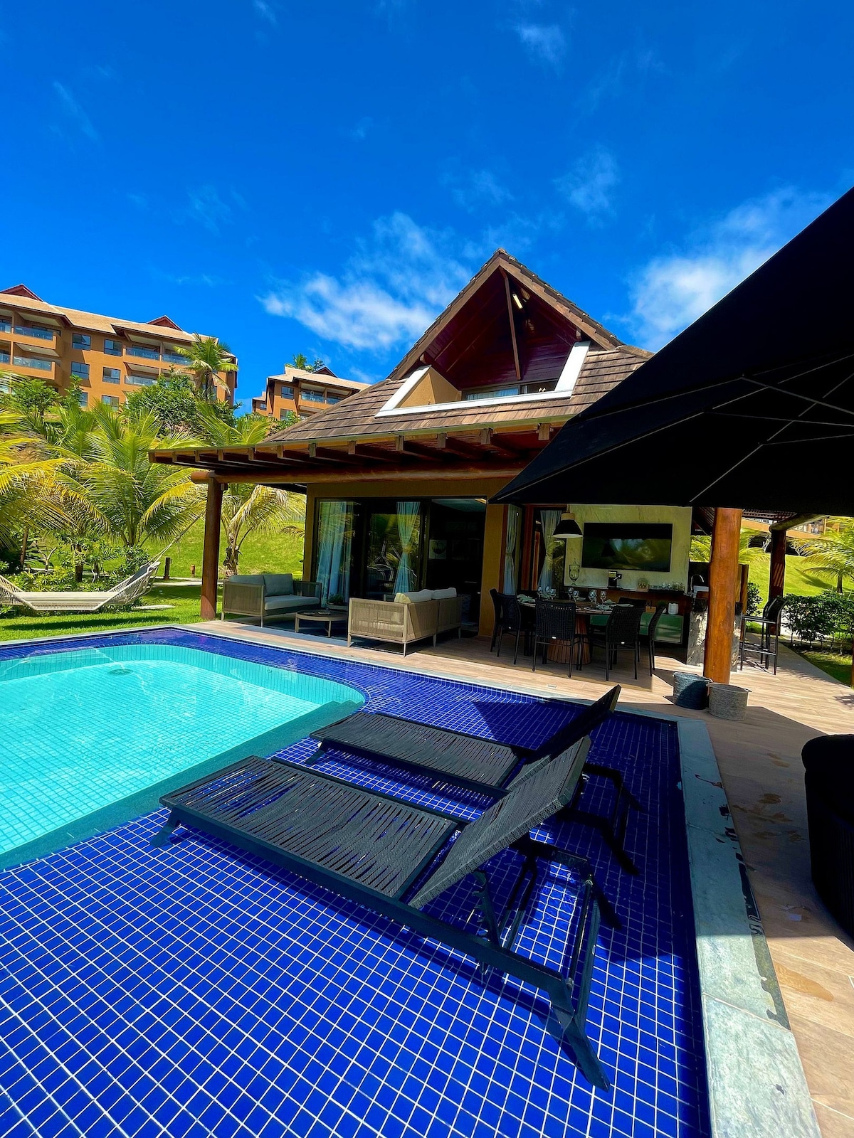 A refreshing swimming pool surrounded by a spacious deck is featured, with two black lounge chairs positioned nearby. The structure beside the pool showcases large glass doors, inviting natural light into the interior. Lush greenery and vibrant blue skies create a peaceful ambiance.