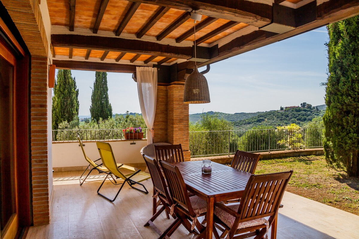 An open terrace furnished with a wooden dining table and chairs is featured. A single lounge chair complements the seating arrangement. The view includes rolling hills and greenery, with a soft blue sky above. Natural light fills the space, enhancing the inviting atmosphere.
