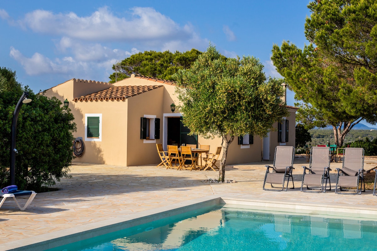 The exterior of the chalet features a spacious outdoor area with a swimming pool in the foreground. Sun loungers are arranged near the pool, while a wooden dining table and chairs are set under a tree. The single-story building showcases light-colored walls and several windows.
