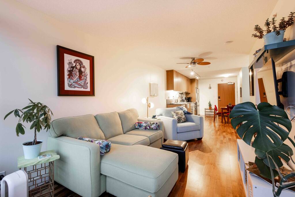A modern living area features a comfortable L-shaped sofa with colorful cushions and a side table adorned with a plant. Light wooden flooring contrasts with the bright walls, leading to a dining space in the background. A ceiling fan and recessed lighting enhance the open atmosphere.