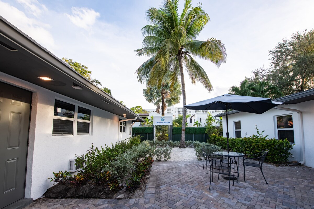 An outdoor area is presented, featuring a brick-paved patio surrounded by lush greenery. A round table with four chairs sits under a shaded umbrella, offering a comfortable spot for relaxation. Towering palm trees frame the space, contributing to the serene atmosphere.
