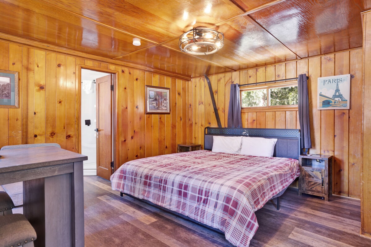 A spacious sleeping area is surrounded by warm wood paneling, featuring a large bed dressed in plaid bedding. Two windows allow natural light to brighten the room. A doorway leads to the bathroom, while a small dining area with stools is visible in the corner.