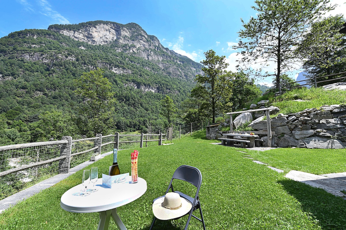 A spacious outdoor area is presented, featuring a small white table set with glasses and a bottle, overlooking lush green grass. A folding chair is positioned nearby. The scenic backdrop showcases towering mountains and a clear blue sky, framed by trees and a stone fence.