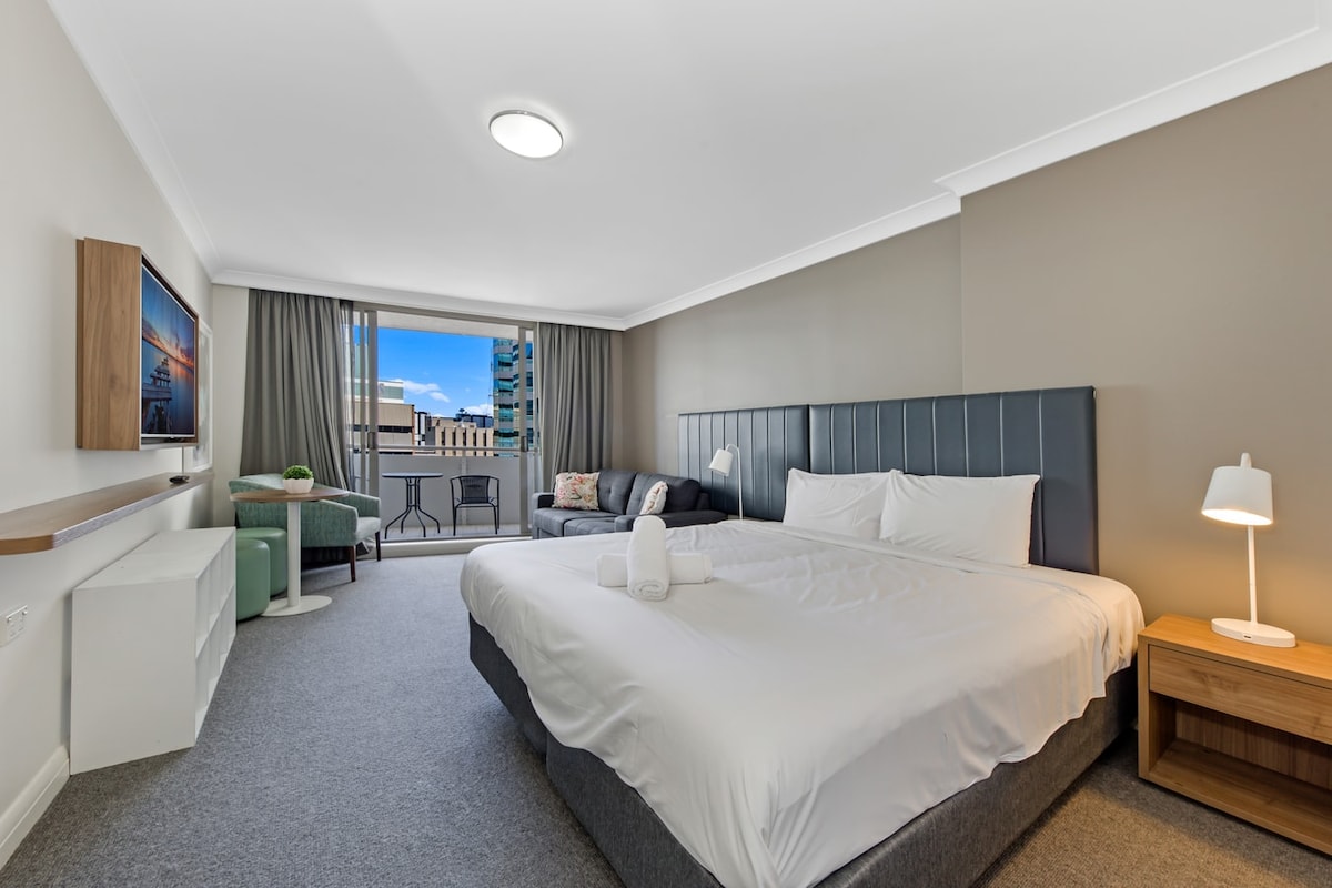 A spacious studio features a king bed with two rolled towels placed neatly on the covers. A double-size sofa bed is positioned nearby, alongside a small armchair. Natural light enters through the balcony doors, with a view of the outside cityscape visible from the window.