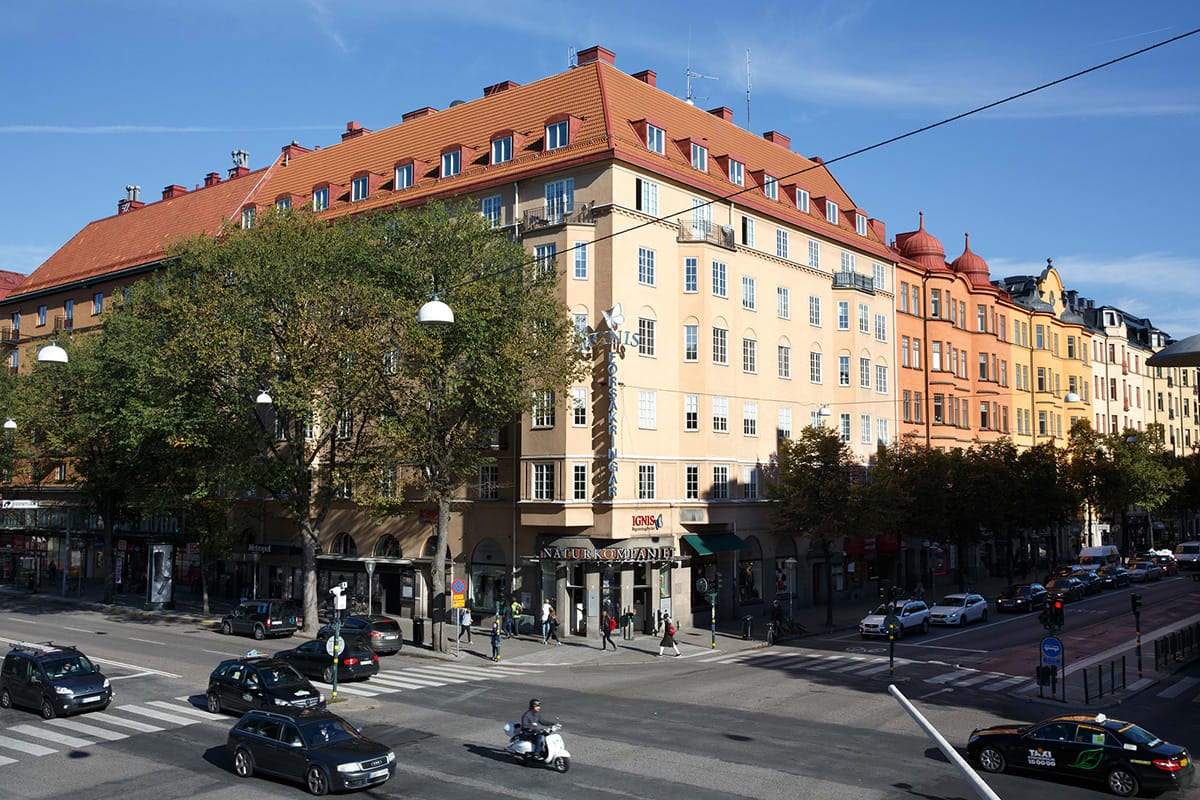 A bustling street scene captures the vibrant urban setting of Vasastan, with a mix of vehicle traffic and pedestrians. The foreground features a crosswalk, while the background showcases a colorful apartment building with characteristic red roofs and numerous windows, framed by lush green trees.