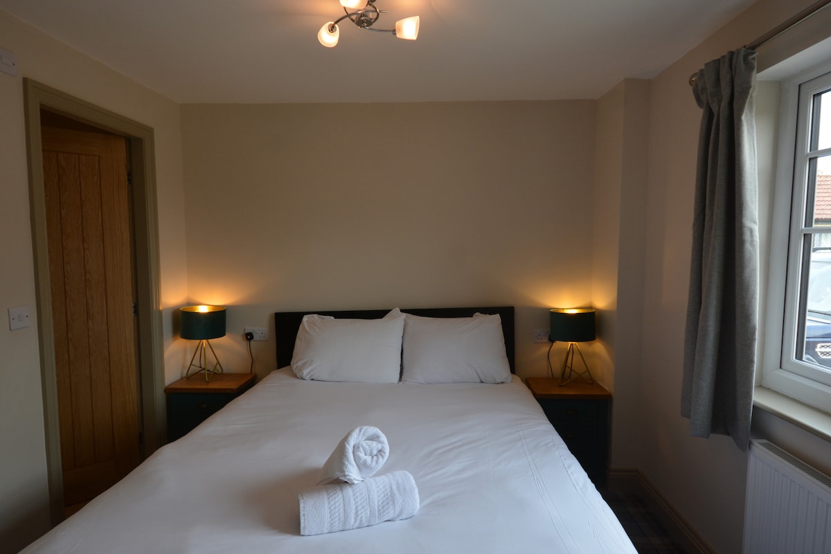 A welcoming bedroom features a spacious bed with plush white linens, flanked by bedside tables and stylish lamps. Soft, neutral walls create a calming environment. Light enters through a window dressed with light curtains, enhancing the room's cozy feel.