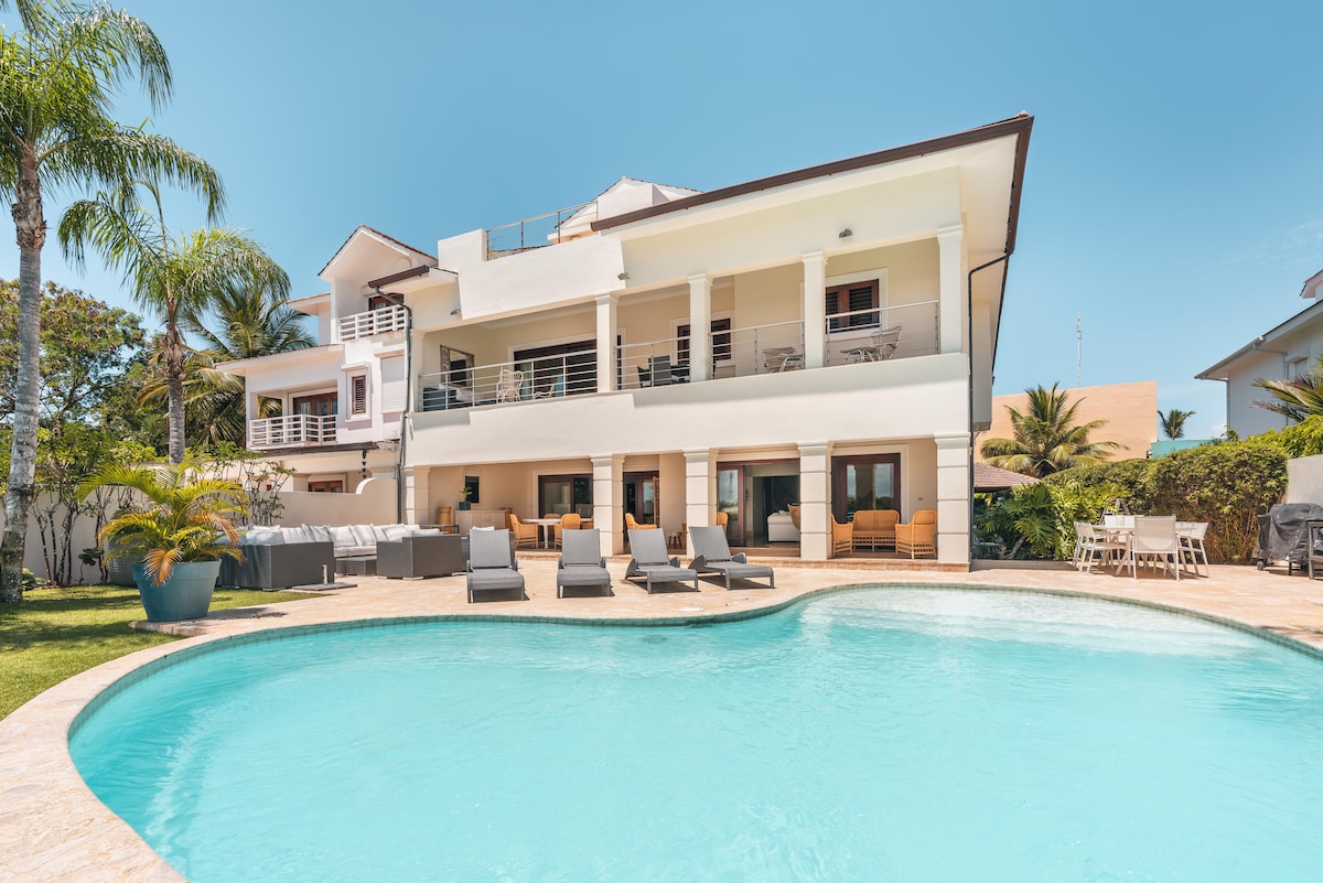 A spacious villa is showcased with a serene swimming pool in the foreground. Lounge chairs surround the pool area, while the two-story structure features a large balcony with seating. Lush greenery is present, complementing the sunlit exterior.
