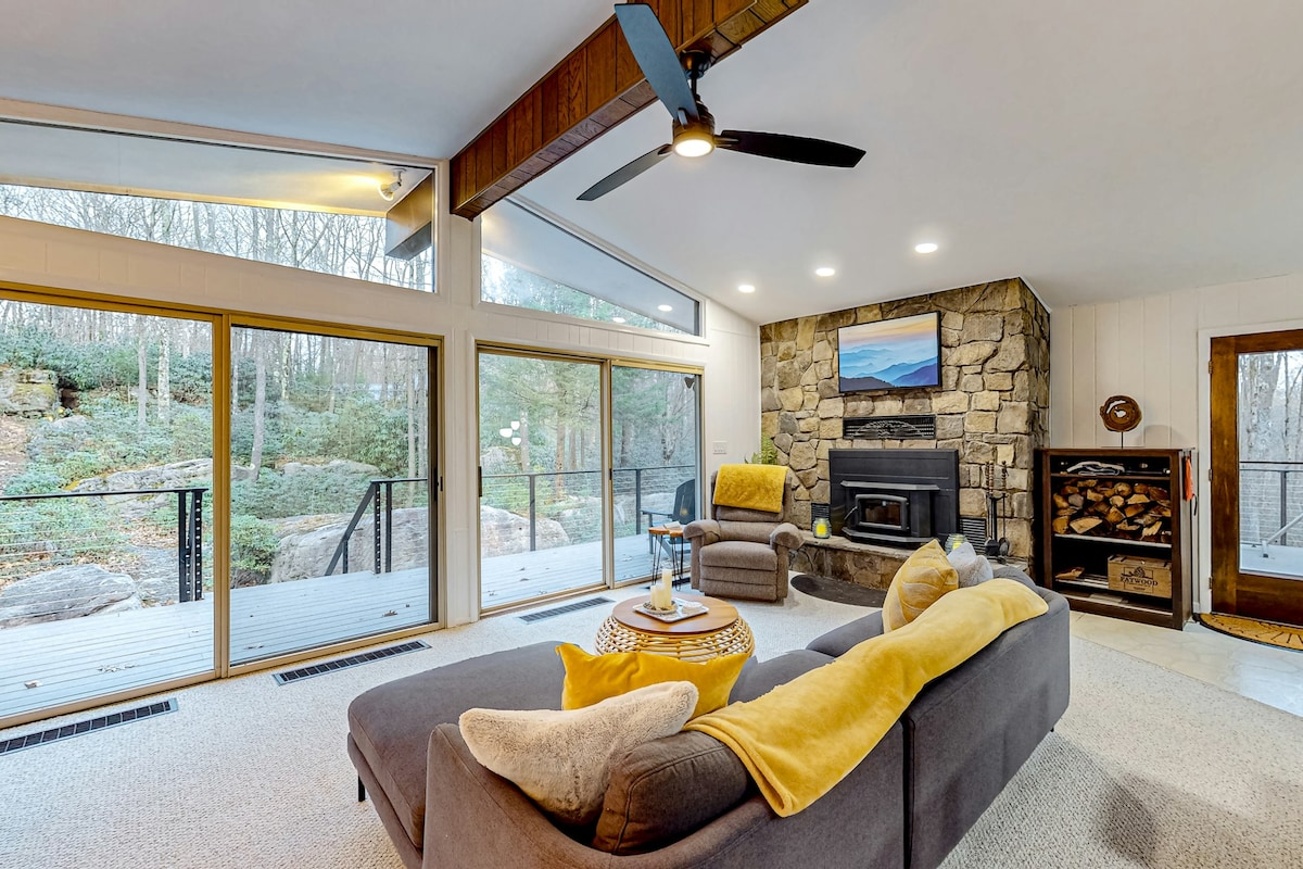 A spacious living area features vaulted ceilings and floor-to-ceiling windows, providing views of the surrounding nature. A cozy wood-burning fireplace is set against a stone wall. Comfortable seating is arranged around a coffee table, while natural light brightens the carpeted space.