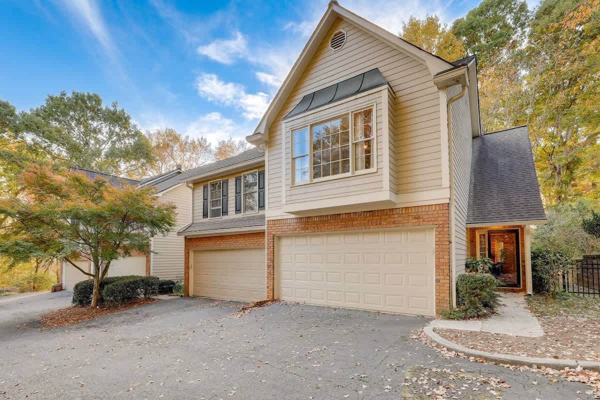 A spacious two-car garage is visible in this well-maintained townhome exterior. The building presents a mix of natural brick and soft beige cladding. Lush greenery surrounds the entrance, with seasonal foliage adding color to the scene under a clear blue sky.