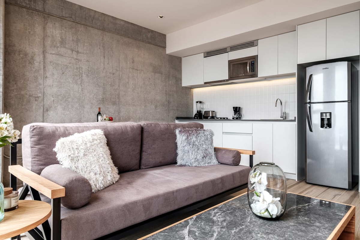 A modern living area features a plush gray sofa adorned with soft throw pillows. A sleek kitchen with white cabinetry and stainless steel appliances is visible in the background. A glass coffee table with a decorative vase sits in front of the seating area.