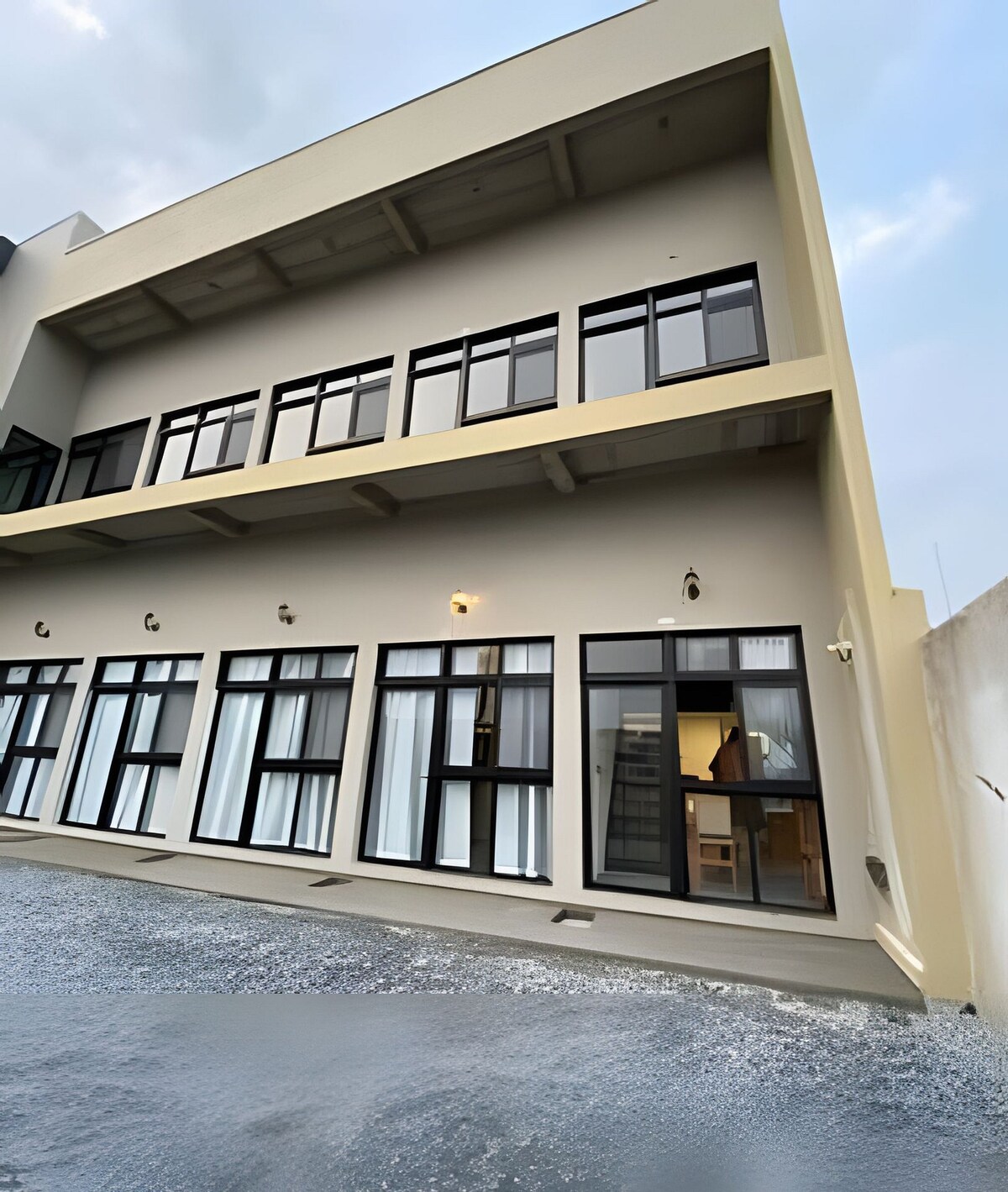 The exterior of the building features a modern design with multiple floor-to-ceiling windows, providing ample natural light. A smooth surface extends before the structure, while steps lead to the welcoming entrance. The contemporary façade is complemented by neutral tones and simple light fixtures.