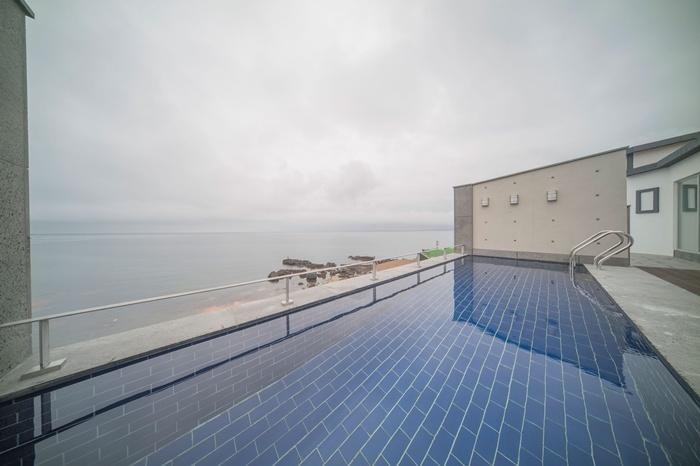 An outdoor swimming pool with a tiled surface reflects the sky, set against a backdrop of calm waters. The minimalist architecture of the surrounding area enhances the serene atmosphere, while the faint outline of rocks can be seen along the shore in the distance.