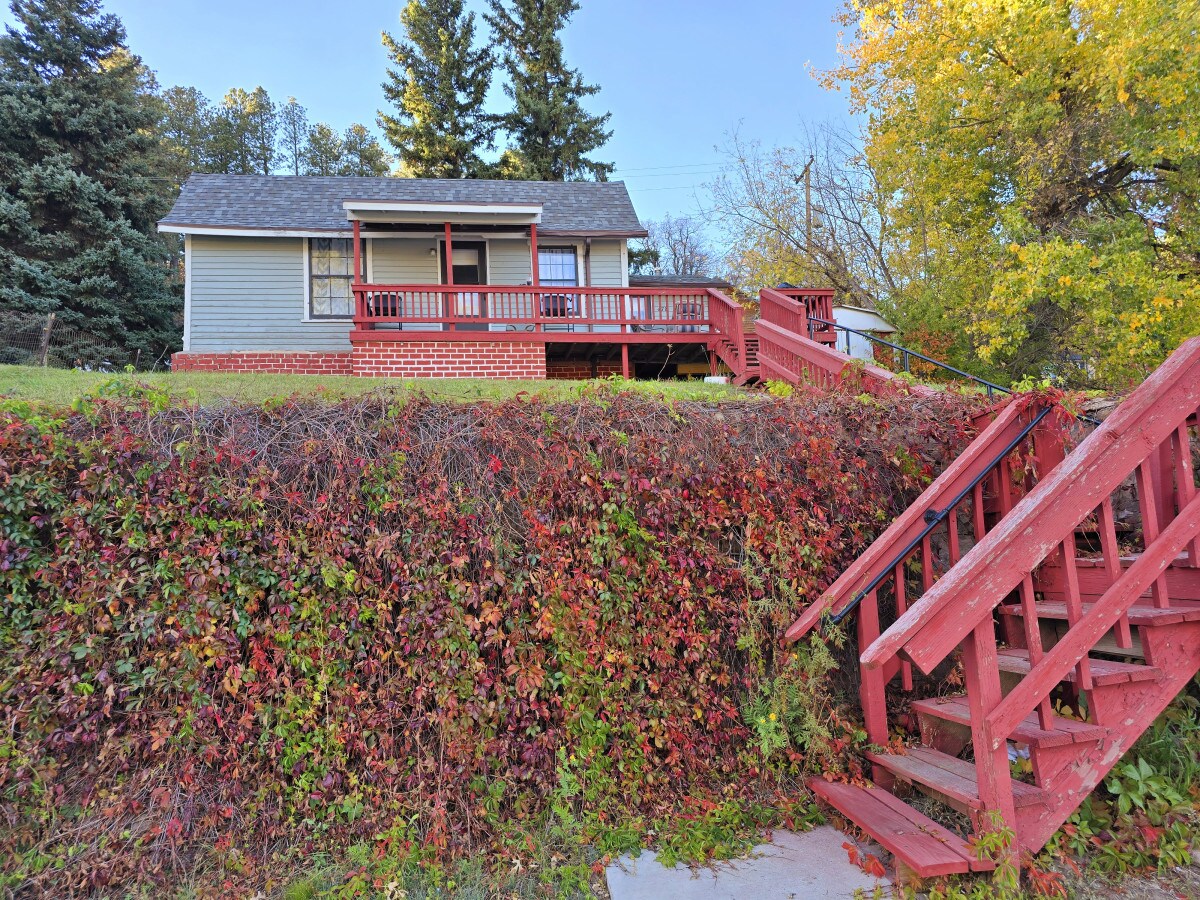 A charming bungalow is set on a slight elevation, featuring a red wooden staircase leading to the front porch. Lush greenery and vibrant climbing plants frame the base of the house, while large windows invite natural light into the inviting space.