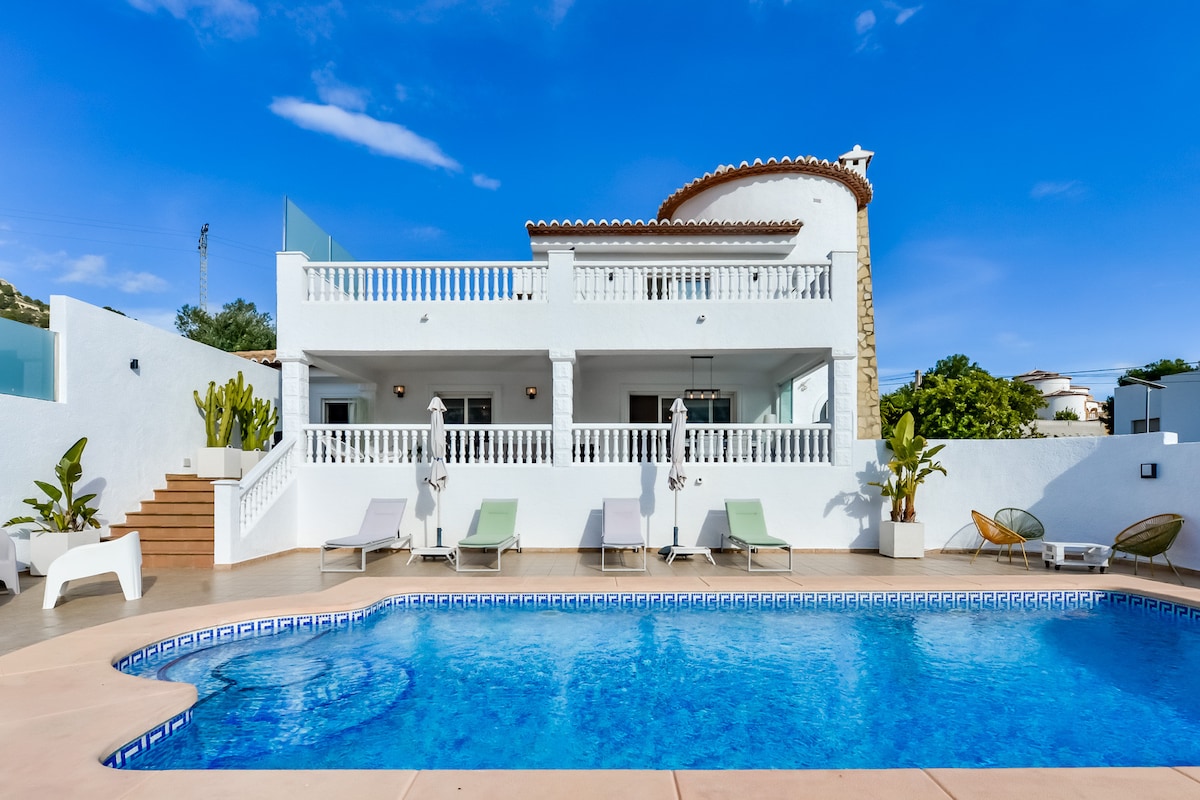 A spacious villa is depicted with a large swimming pool at the forefront, surrounded by sun loungers. The two-story structure features a white facade, with a terrace visible on the upper level. Lush greenery is present, contributing to a tranquil outdoor space.