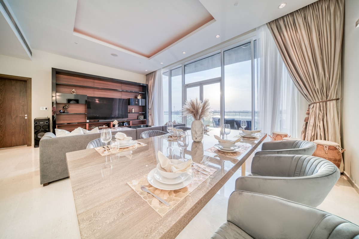 The dining area features a spacious table set for multiple guests, surrounded by stylish chairs. Large windows allow natural light to fill the space, showcasing views of the outside. Elegant decor elements, including a modern TV unit and soft drapes, complement the inviting atmosphere.