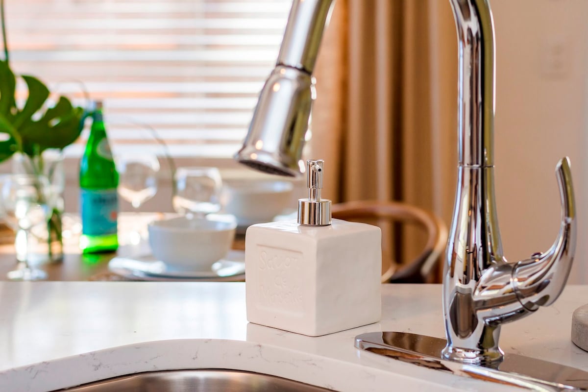 A modern kitchen sink area features a polished chrome faucet beside a white soap dispenser. In the background, glassware and tableware are visible, accompanied by green and clear bottles, enhancing the functional kitchen space.