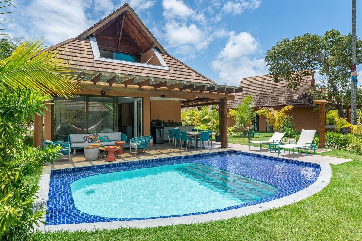 A modern villa is framed by green landscaping, featuring a private pool with a blue tiled edge. Outdoor seating is arranged near the pool, alongside a dining area under a shaded patio. The home’s architecture includes a steep roof and large windows for natural light.
