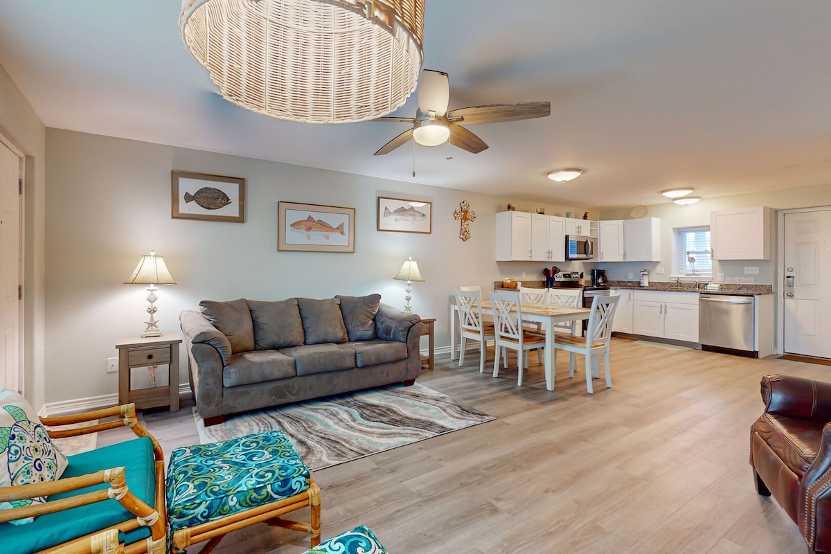 A spacious living and dining area features a comfortable sofa and armchairs, with decorative cushions adding a pop of color. A dining table for six is set beside a modern kitchen equipped with stainless steel appliances and white cabinetry. Light fixtures provide ample illumination throughout the space.