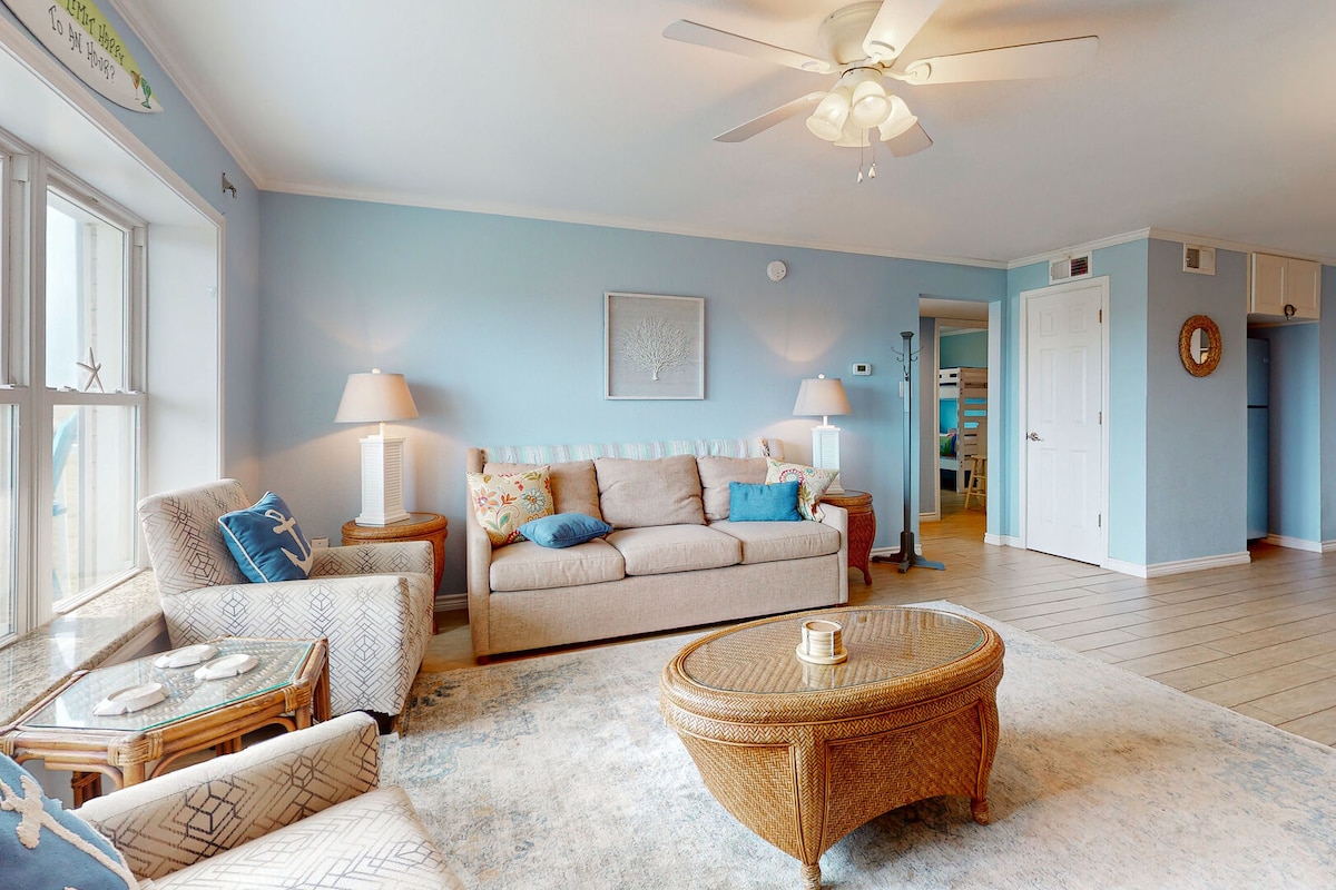 The living area is adorned with light blue walls and features a spacious sofa, a wicker coffee table, and two lamps beside the windows. Natural light fills the room, enhancing the airy atmosphere. Open access leads to a kitchen area in the background.