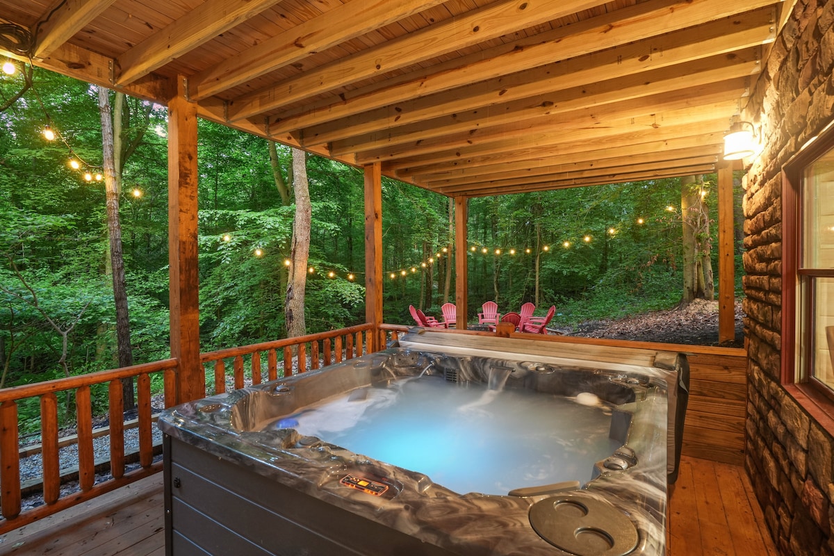 A hot tub is positioned on a covered porch, surrounded by a tranquil wooded landscape. Soft blue lighting emanates from the hot tub, creating a relaxing atmosphere. String lights hang above, illuminating the area, while outdoor seating is provided by bright pink chairs in the background.