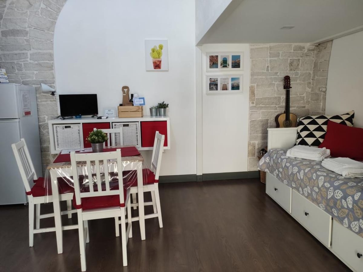 The open living area features a kitchen enclosed by a white stone arch. A dining table with four white chairs is positioned beside a red sideboard and a TV. A cozy seating area includes patterned cushions, with white towels neatly arranged on a bed in the background. Wall art decorates the space.