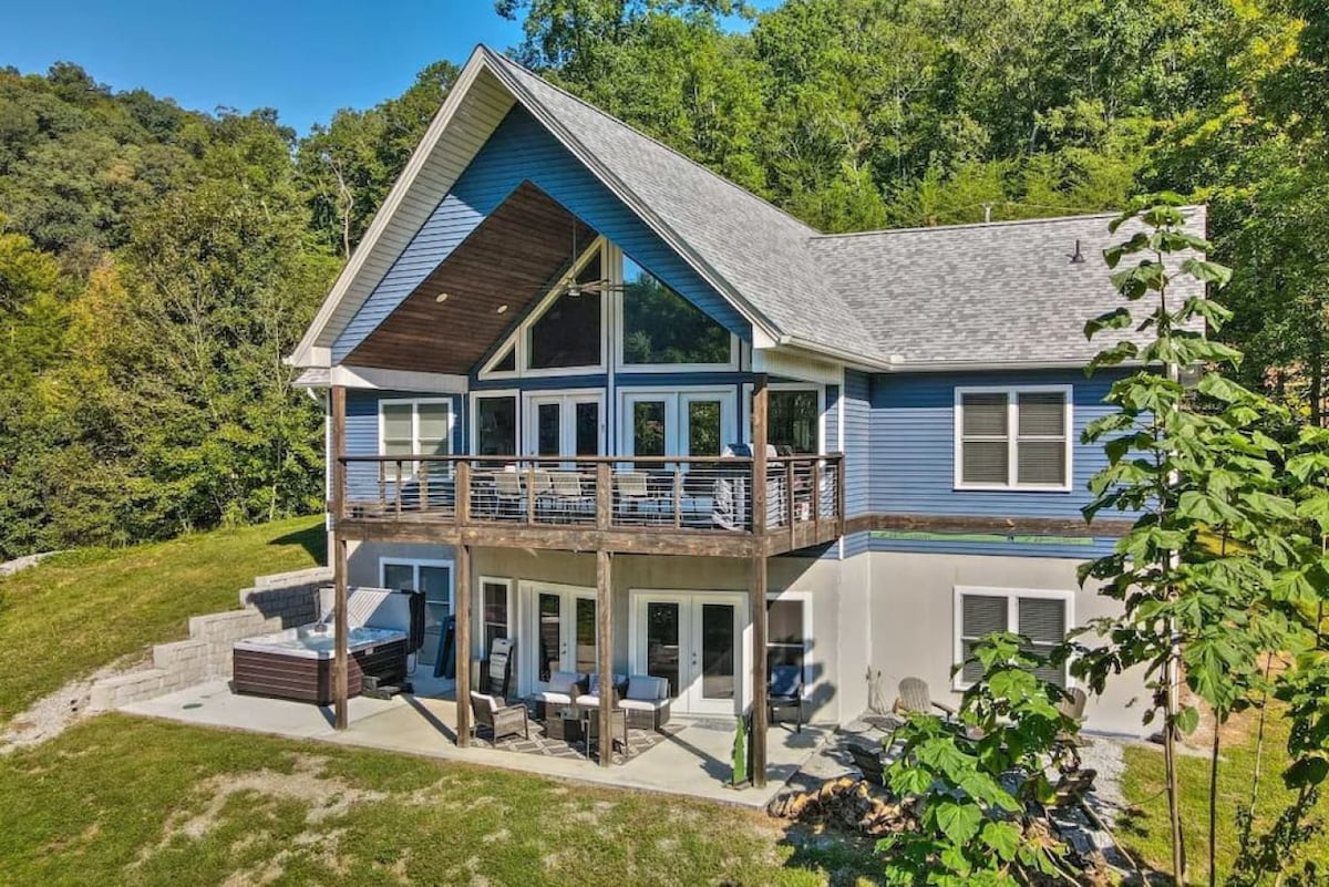 The exterior of a modern lakefront home is framed by lush greenery. A spacious deck features seating areas and overlooks the yard. Large windows provide ample natural light, and a hot tub is visible on the deck, inviting relaxation.