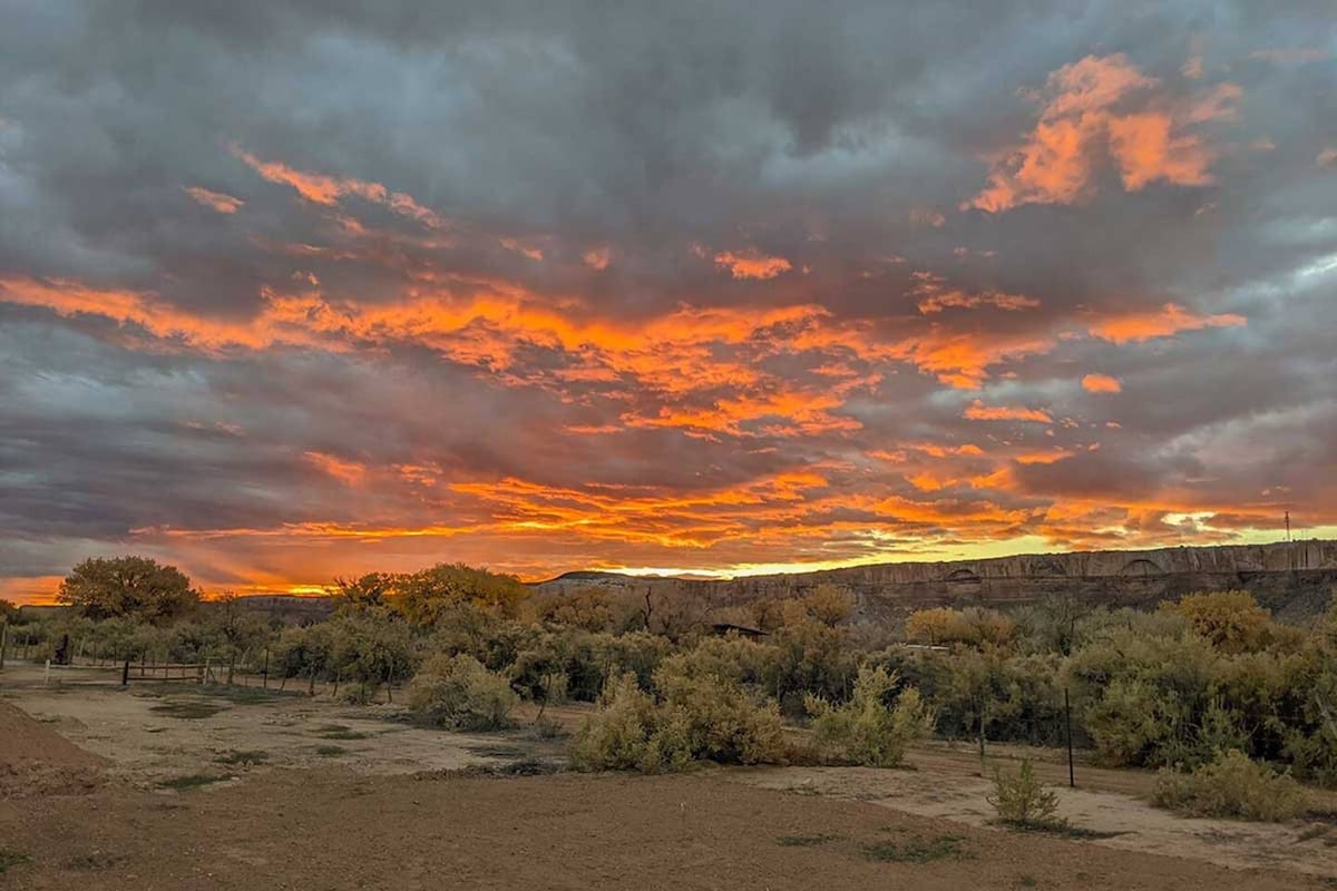 A vibrant sunset casts warm hues of orange and yellow across the sky, contrasting against dark gray clouds. The landscape below features lush, low-lying vegetation and a rocky backdrop, suggesting a natural setting typical of southeastern Utah.