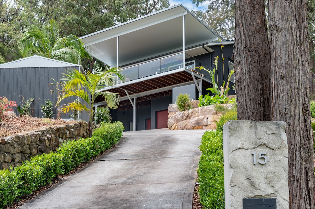 The exterior of the holiday home is presented, showcasing a modern structure set against a backdrop of greenery. A sloped driveway leads to the entrance, bordered by well-maintained landscaping, including a stone wall and palm trees, creating an inviting approach.