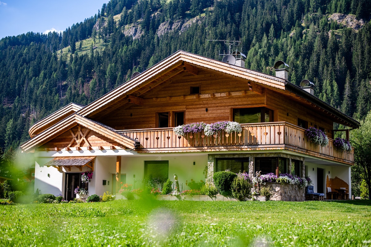 The exterior of the holiday apartment is framed by lush greenery, featuring a wooden structure with decorative balconies adorned with blooming flowers. The design blends harmoniously with the surrounding mountains, showcasing a spacious outdoor area that invites guests to enjoy the natural beauty.