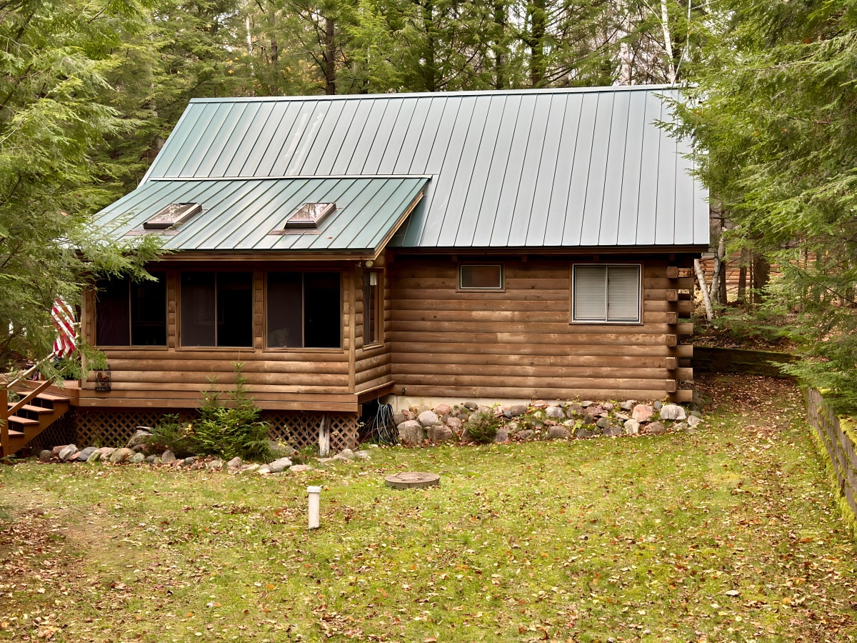 A log cabin features a metal roof and is surrounded by trees and a grassy area. A small wooden porch extends from the front, with a few steps leading down to the yard. Windows are visible, allowing natural light into the cozy structure.