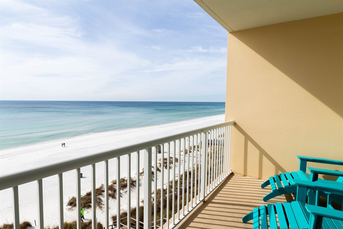 A private balcony features turquoise chairs facing the beach, offering an unobstructed view of the calm turquoise waters and white sandy shores. The horizon stretches into the distance, where the sky meets the sea, enhancing the tranquil coastal atmosphere.