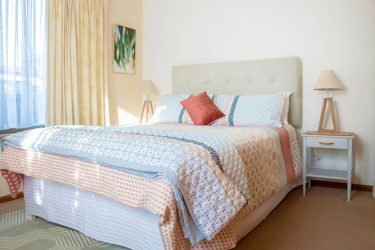 A bright bedroom features a spacious bed dressed in patterned linens. Natural light filters through the sheer curtains, highlighting the soft hues of the decor. Two bedside lamps are positioned on either side, complementing the calm ambiance. A piece of artwork is displayed on the wall.