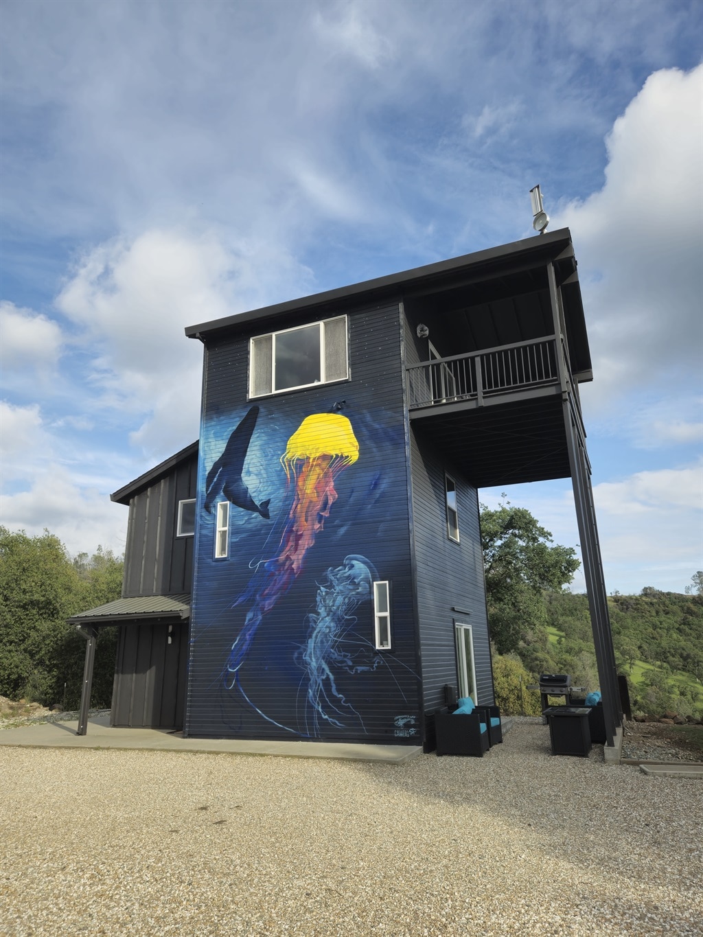 The exterior of the three-story lookout tower features a striking mural with a whale and jellyfish, adding character to the dark blue siding. Surrounding the building, a natural landscape of rolling hills and trees is visible, enhancing the tranquil atmosphere.