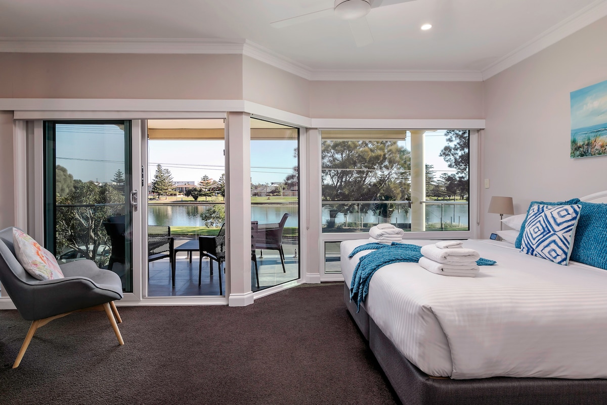A serene bedroom features a spacious bed with white linens and decorative pillows. Large glass doors provide a view of the waterfront, complemented by a small outdoor seating area. A comfortable chair and folded towels are arranged neatly, creating a fresh and welcoming ambiance.