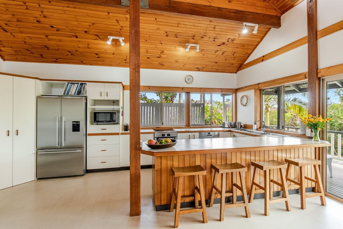 A spacious kitchen features a large island with seating for four and a stainless steel refrigerator. Light filters through expansive windows, illuminating wooden cabinetry and countertops. A microwave and oven are integrated into the design, accented by fresh flowers on the island.
