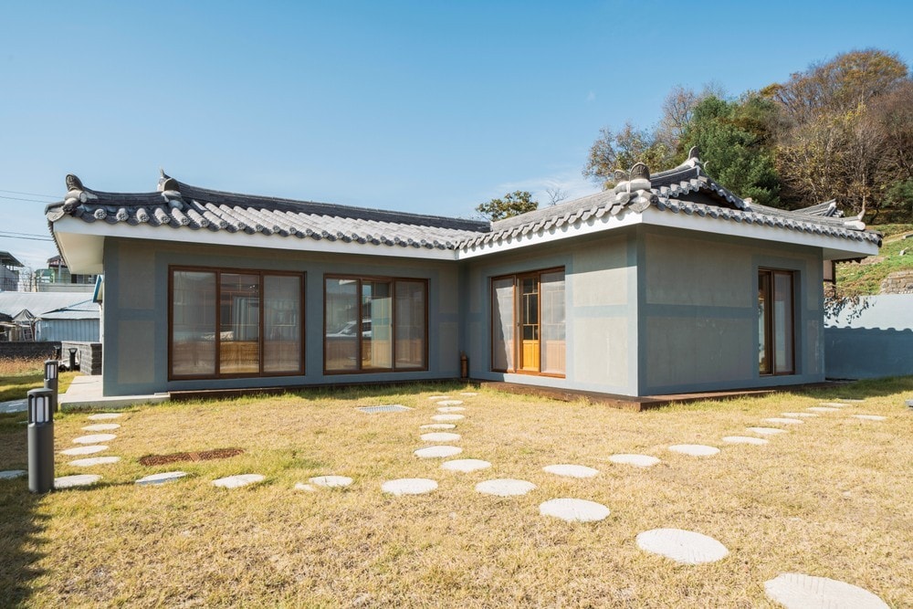 A traditional-style house with a curved tiled roof is surrounded by a manicured lawn. Large windows provide ample natural light, while stepping stones lead through the grassy area, inviting exploration of the outdoor space.