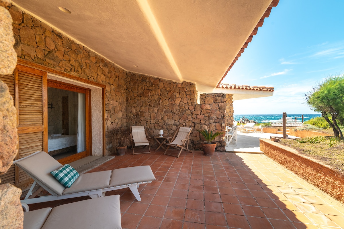 A private outdoor terrace is presented, featuring terracotta tiled flooring and a stone wall. Two loungers are positioned for relaxation, accompanied by a small table. The sea is visible in the background, framed by a clear blue sky, providing an inviting space for guests.