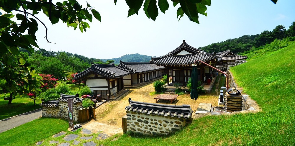 A traditional Hanok structure is surrounded by lush greenery, showcasing a harmonious blend of nature and architecture. The exterior features a thatched roof and wooden beams, with a spacious courtyard inviting relaxation. Pathways lined with stones lead through the serene landscape.