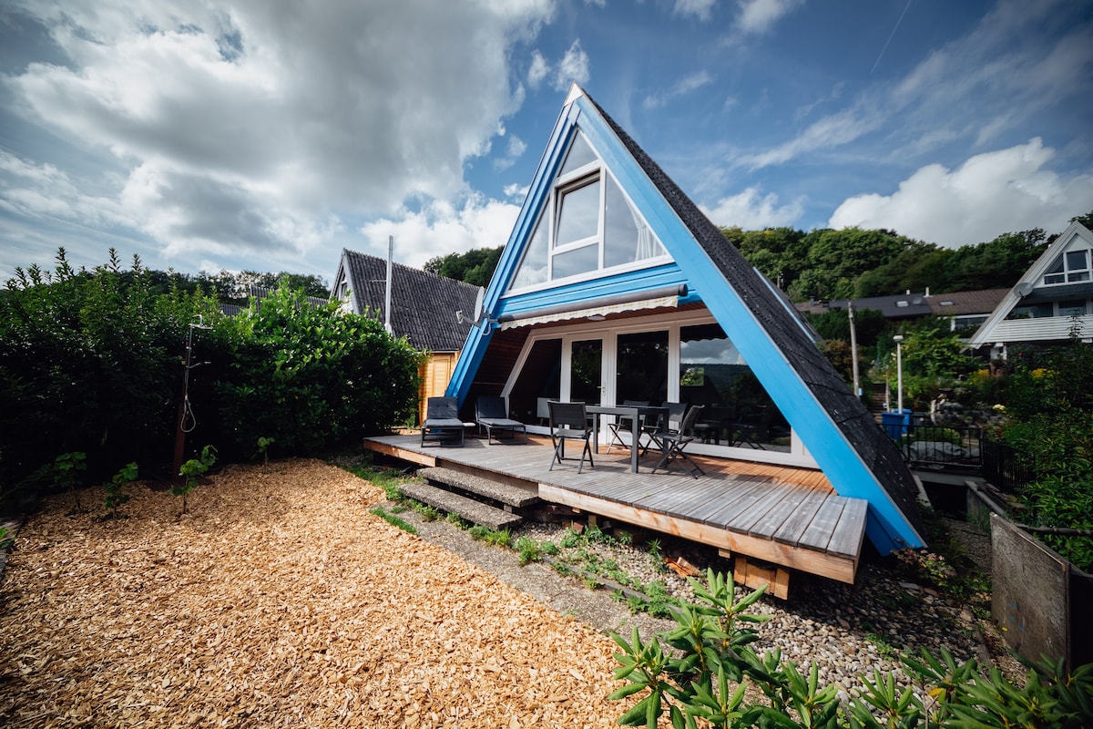 The exterior of a charming A-frame house is showcased, featuring a blue and white façade. A spacious wooden deck with outdoor seating is visible, surrounded by a landscaped area with gravel and greenery. Lush hills are in the background under a partly cloudy sky.