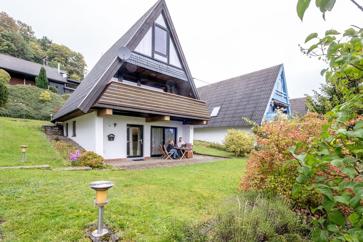 A charming Finn cabin is depicted, surrounded by lush greenery. Two individuals are seated at a small table on the terrace, enjoying the outdoor setting. The structure features a distinctive triangular roof and expansive windows that invite natural light.