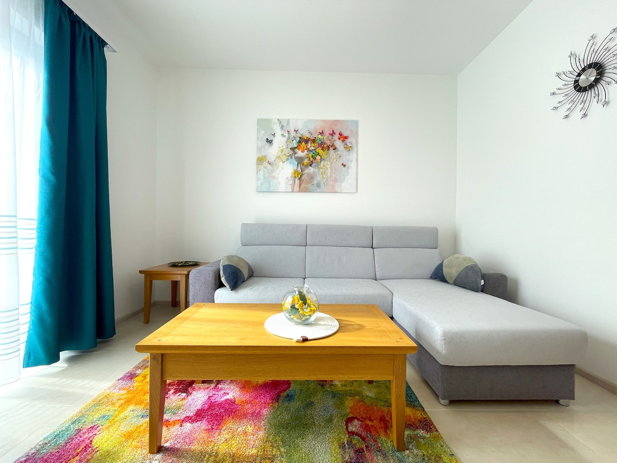 Pumzika katika eneo la kisasa la kuishi na sofa ya sehemu ya kijivu, zulia lenye rangi nyingi, meza ya kahawa ya kifahari na michoro midogo. Chumba hiki chenye hewa safi ni kizuri kwa ajili ya kupumzika baada ya kuchunguza jiji.