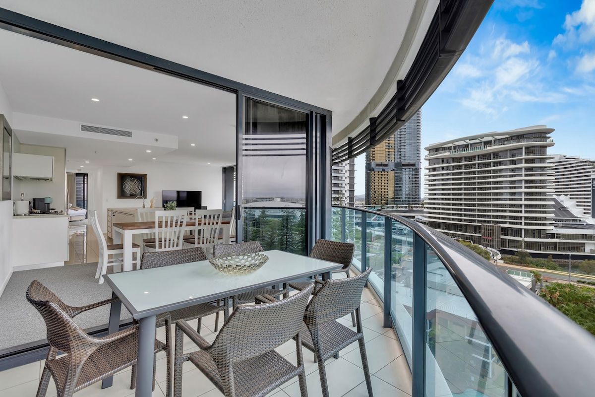 A spacious balcony features a glass table surrounded by wicker chairs, offering a setting for outdoor dining. The scene includes views of the cityscape, with modern buildings and greenery visible in the background. Natural light fills the area through large sliding doors.